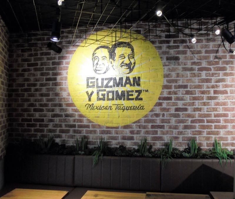 Painted Guzman y Gomez logo on a recycled-brick feature wall above banquette seating, from a C&A fit-out.